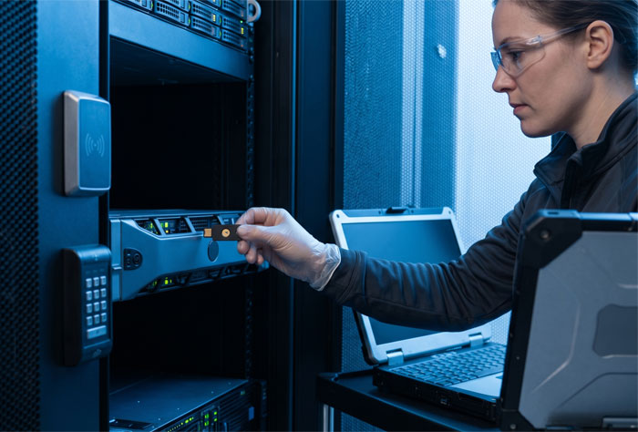 Local serveur sécurisé montre une experte en cybersécurité insérant une clé de sécurité matérielle dans un serveur rack d'entreprise. Elle porte un gant de précision technique et utilise un ordinateur portable de diagnostic posé sur un chariot mobile, illustrant les actions de maintenance continue et de sécurisation de l'actif numérique d'un cabinet.